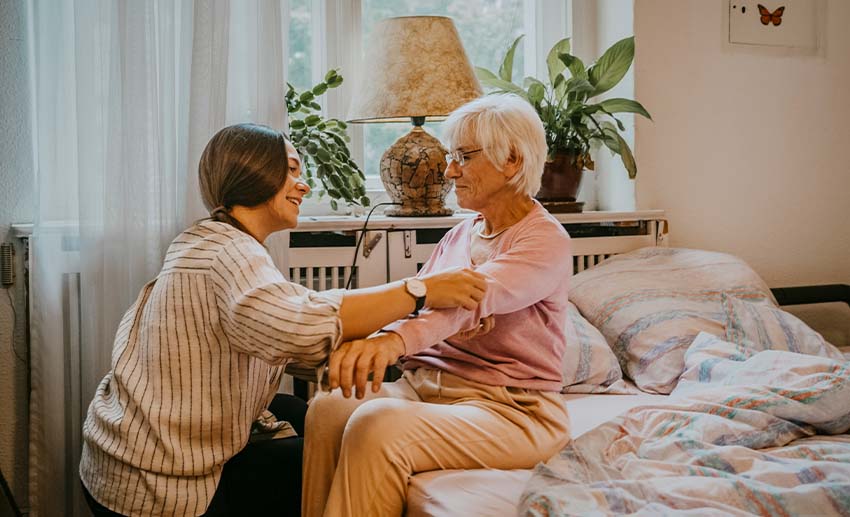 A woman kneels and checks the blood pressure of an older person sitting on a bed