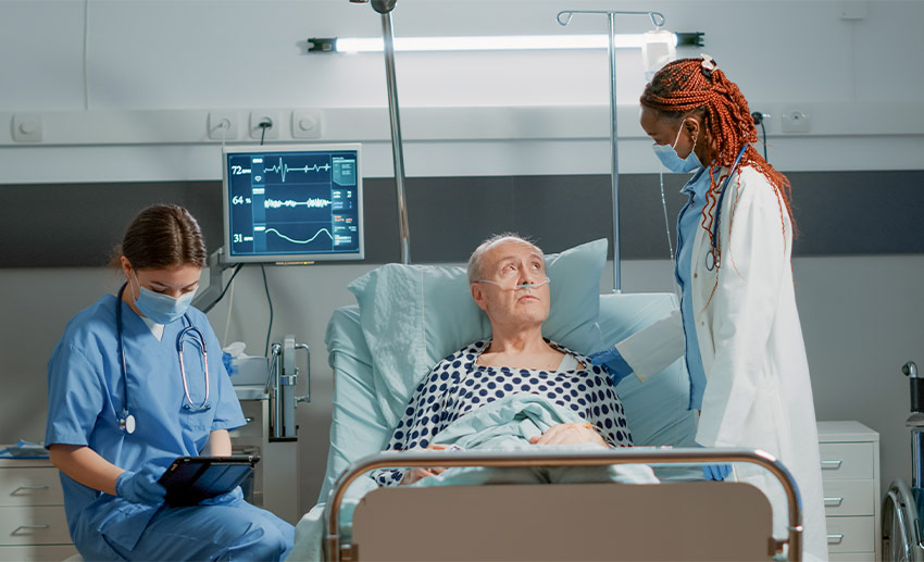 A patient in a hospital bed with a doctor standing next to them and a nurse seated in a bedside chair