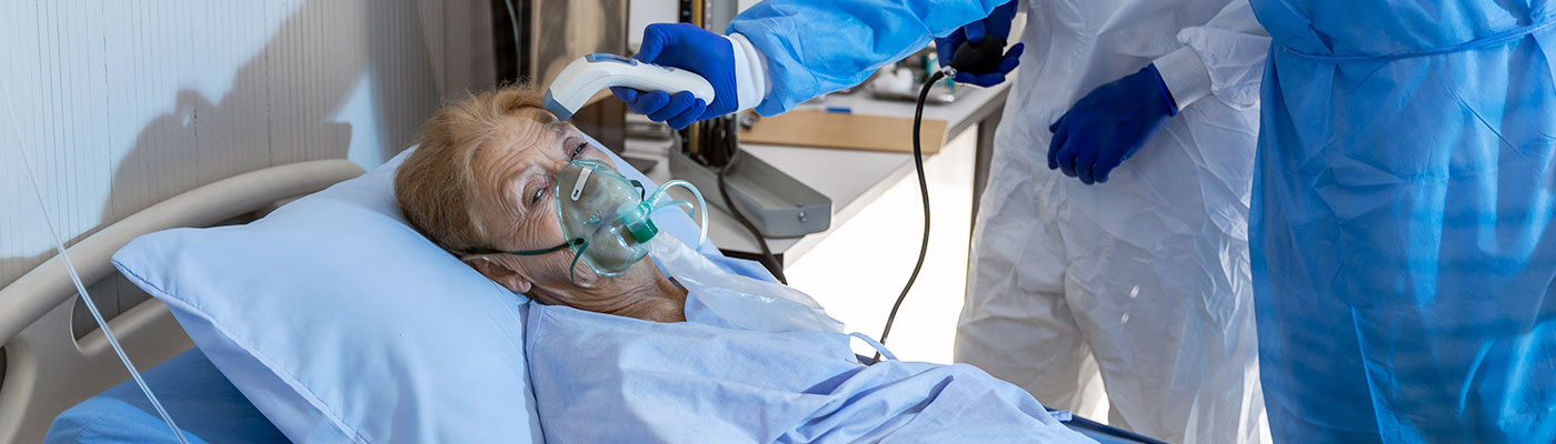 A healthcare worker takes the temperature of a woman in a hospital bed who wears a mask for supplemental oxygen
