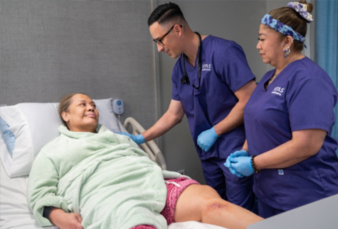 Two nurses in purple scrubs next to a hospital bed with a female patient