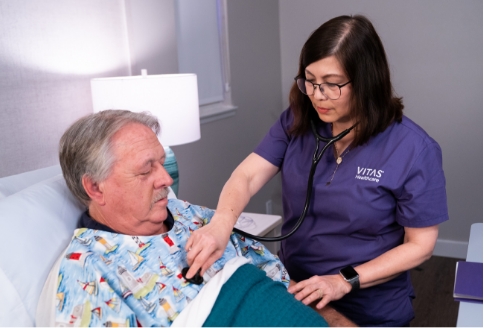 A nurse in purple scrubs checks on a patient in a bed